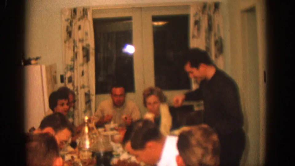 1963 Zurich home-movie frame of adults seated at a dining table as a man in a dark sweater serves or passes food.