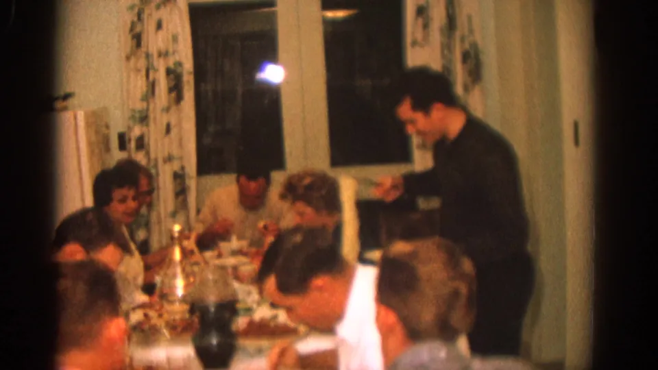 A warm 8mm view of a Zurich family dinner, with several adults eating and one man standing near the table.