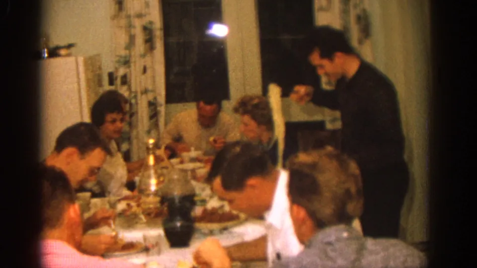 1963 Zurich family meal frame showing diners around a crowded table, patterned curtains, and a man serving from the side.