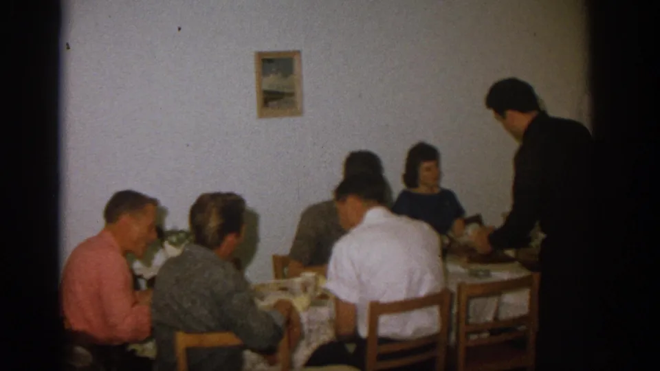 Zurich 1963 home interior with adults eating at a table and a standing man near the right side of the frame.