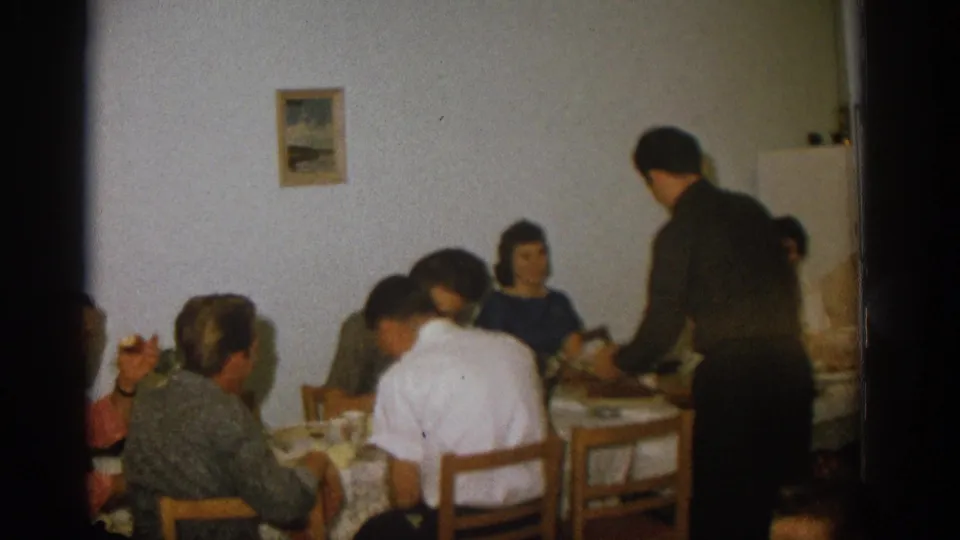 Zurich 1963 home-movie frame of a man standing beside adults seated at a dining table in a plain interior.