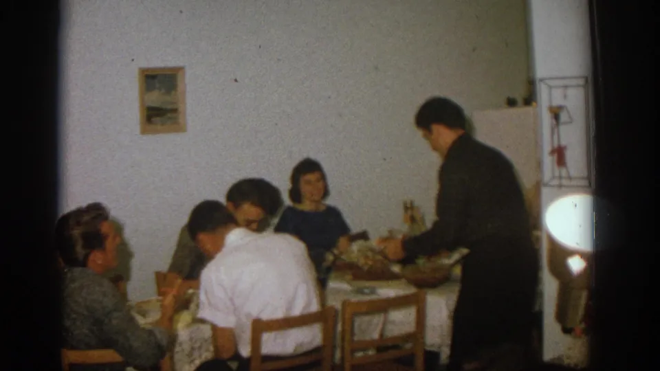 Zurich 1963 home dinner scene with adults seated around a table as a standing man appears to serve food.