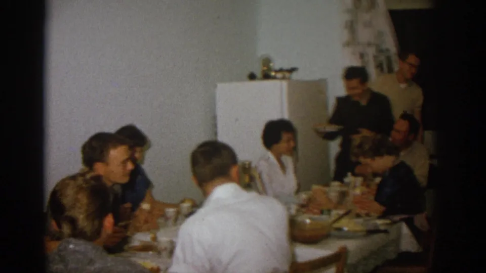 1963 Zurich home-movie frame showing adults gathered around a kitchen dinner table with serving dishes and a white refrigerator.