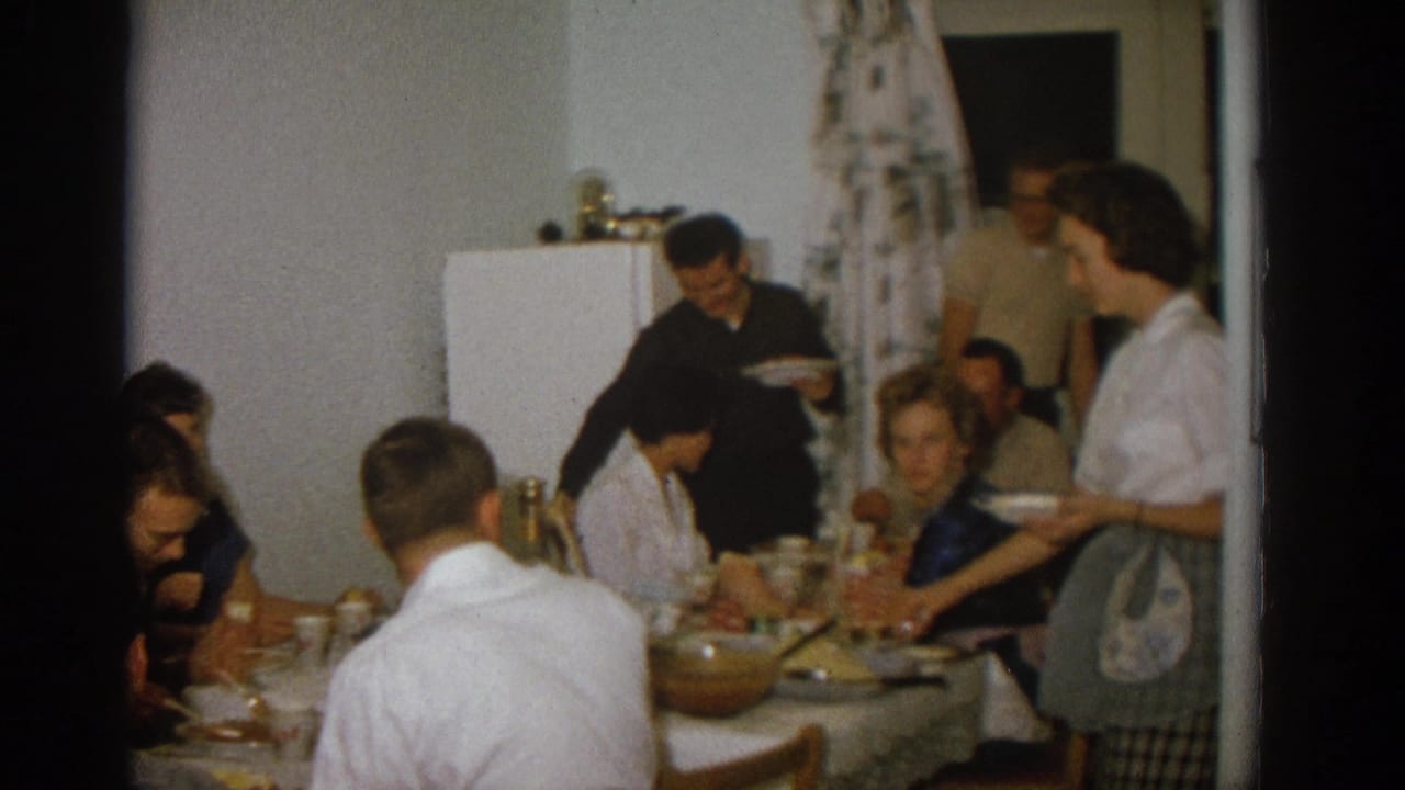 Woman in an apron stands beside the table as diners eat and a man carries a plate.