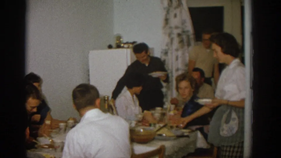 1963 Zurich kitchen dinner scene with an apron-wearing woman serving near seated adults and a man holding a plate.