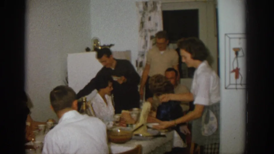 1963 Zurich home-movie frame of adults serving and eating at a crowded kitchen table in a private interior.