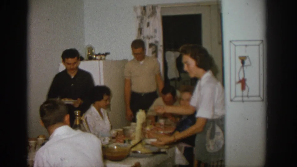 1963 Zurich kitchen dinner frame showing a woman reaching across the table as adults gather around the meal.