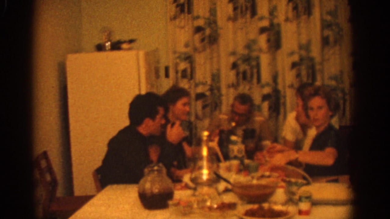 1963 Zurich home-movie frame showing adults gathered at a kitchen dinner table with a white refrigerator and patterned curtains behind them.