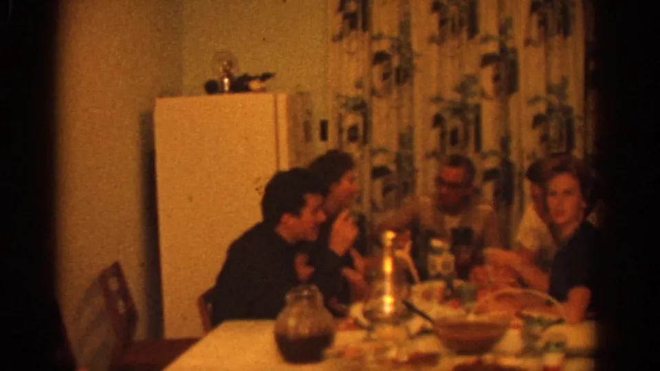 Adults at a Zurich kitchen table in 1963, with meal dishes, glasses, patterned curtains, and a white refrigerator visible in soft 8mm color.