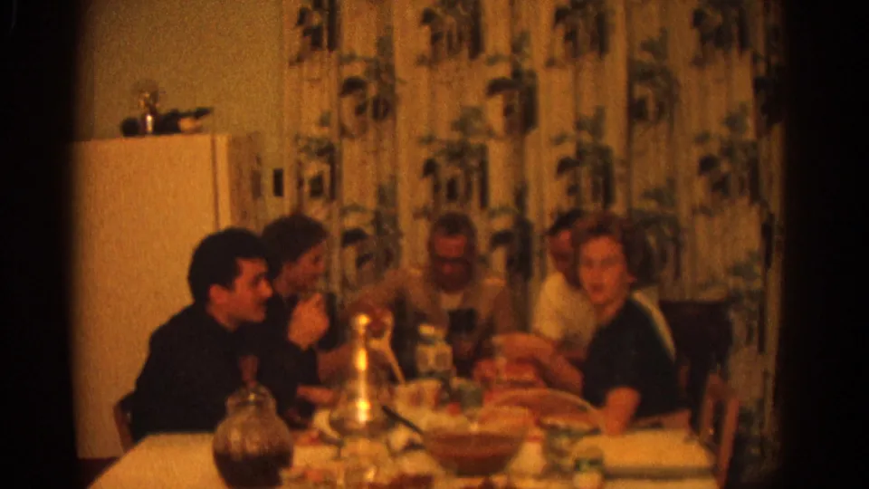 Warm 8mm frame of a 1963 Zurich family dinner, with adults seated at a full table inside a private kitchen.