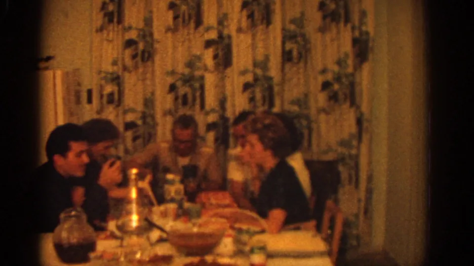 Late moment from the 1963 Zurich dinner scene, with adults leaning in at the table amid dishes, glasses, curtains, and warm indoor light.