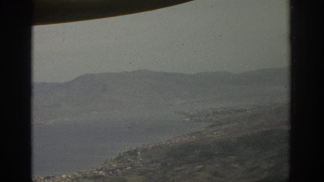 1963 8mm view of a broad Swiss lake, hazy mountain ridges, and shoreline settlement seen through an aircraft window frame.