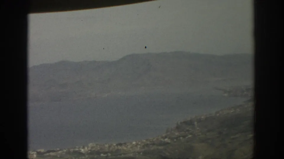 A soft 8mm aerial view shows a lake, green hills, faint shoreline buildings, and distant mountains under a pale sky.