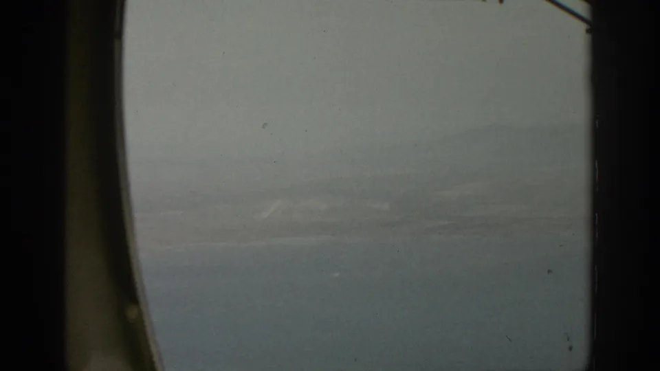 1963 home-movie aerial view of a lake or broad water surface with pale shoreline shapes below haze.
