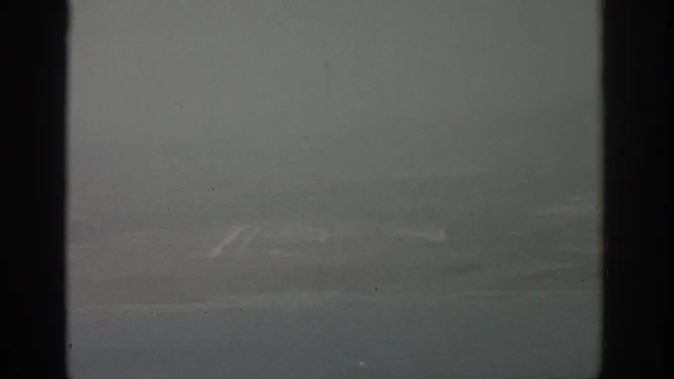 Aircraft-window view near Zurich with hazy water, a faint shore, and distant hills on 1963 8mm film.