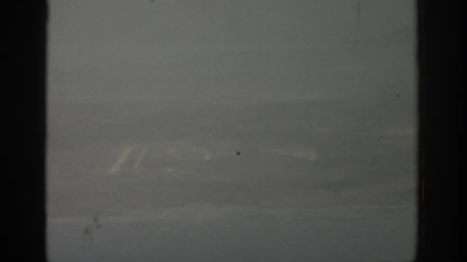 1963 home-movie frame showing muted terrain through an airplane window, softened by haze, grain, and scan texture.