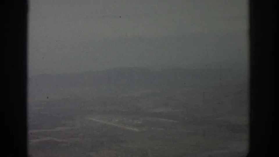 A 1963 8mm airplane-window view near Zurich with faint ground patterns, haze, and dark window borders.