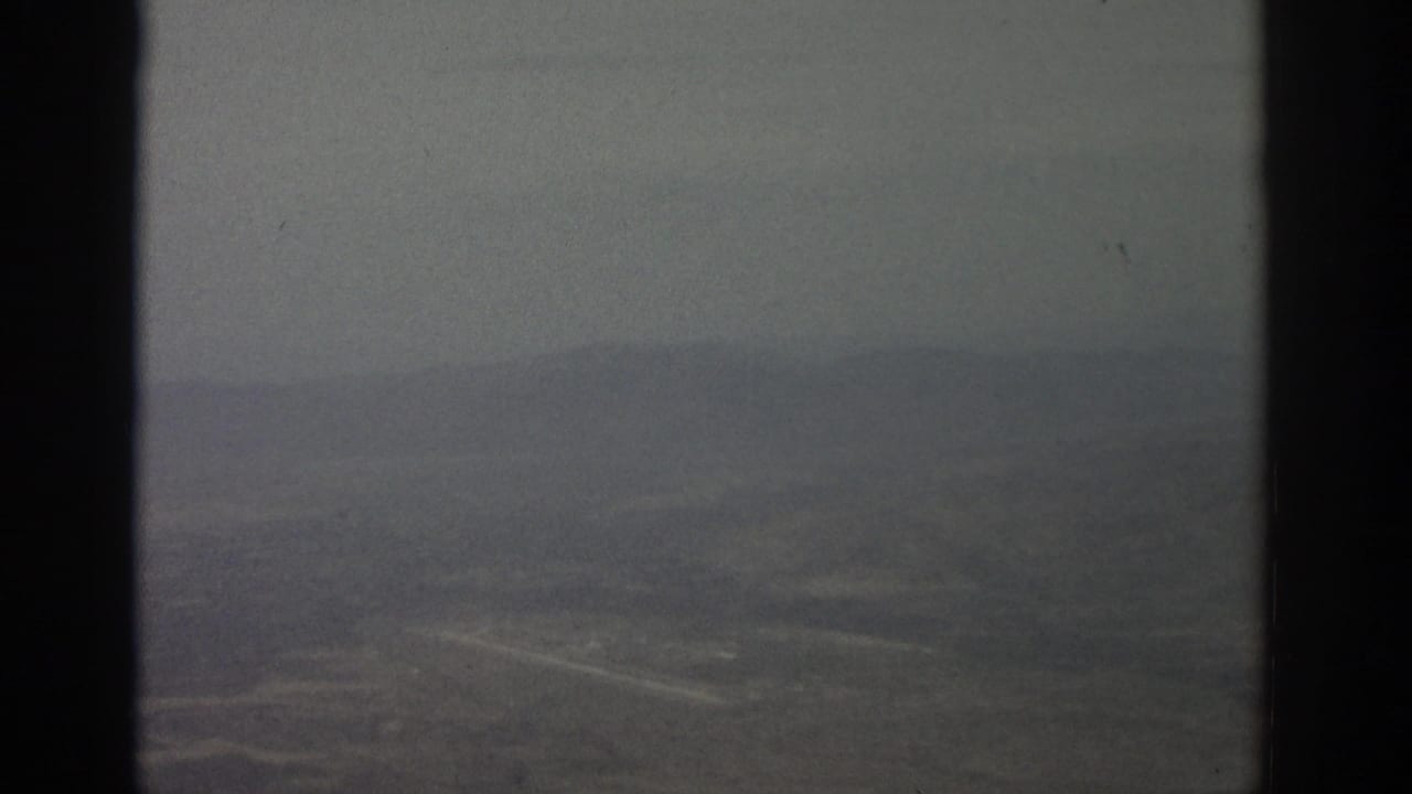 1963 8mm-style airplane-window view over a hazy landscape with faint roads and dark vertical frame borders.