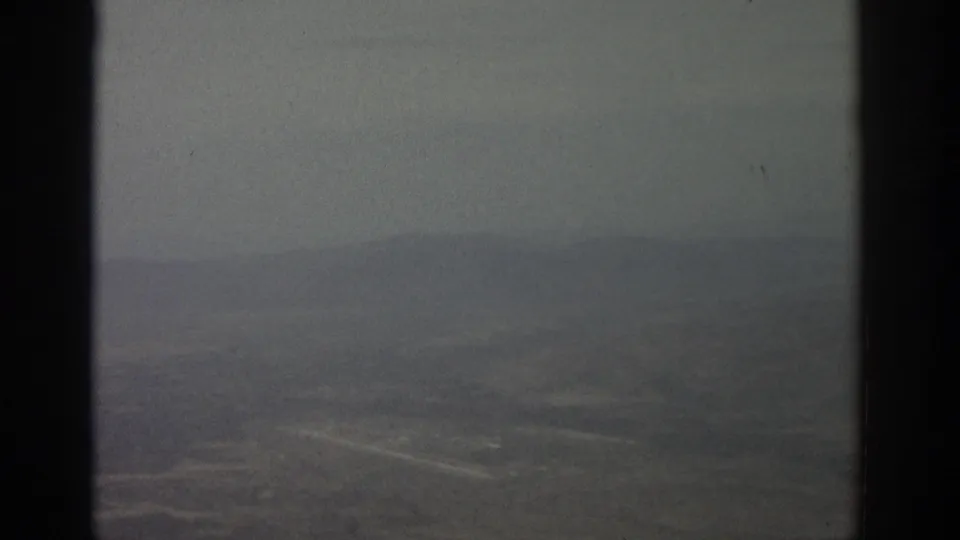 1963 8mm-style airplane-window view over a hazy landscape with faint roads and dark vertical frame borders.