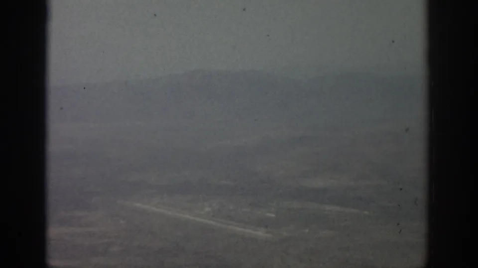 Airborne 8mm frame showing muted terrain below, a pale road line, and dark airplane-window dividers.