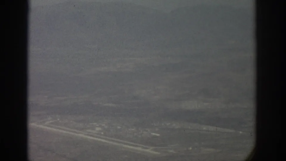Small-gauge film frame of a hazy aerial landscape, with a bright road or runway-like line and dark aircraft-window borders.