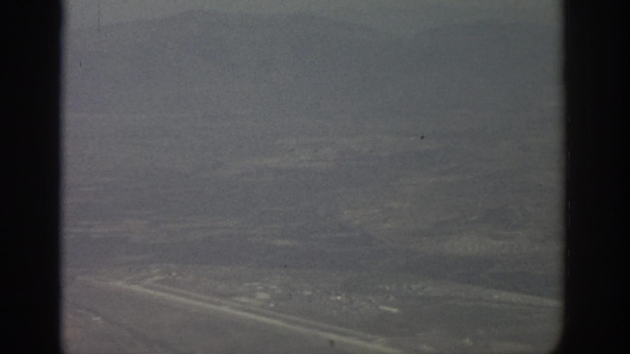 1963 Zurich-area aerial view through a dark airplane window frame, showing hazy open land and distant city texture.