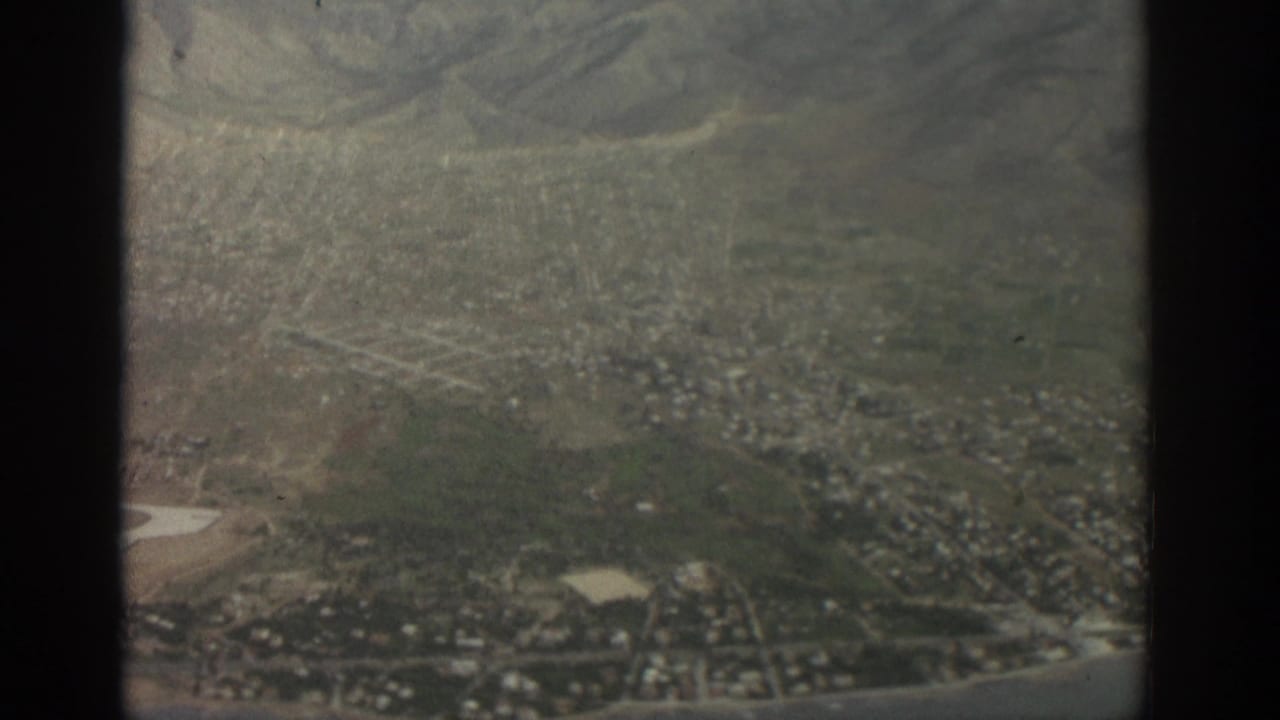 Aerial view of city blocks, hills, and an airfield seen through a window frame.