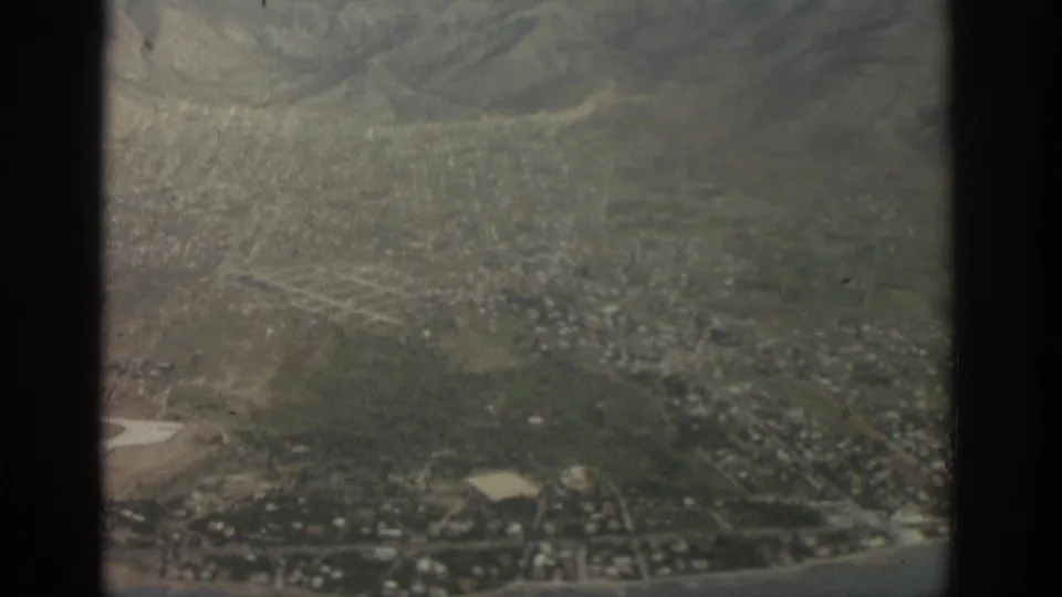 1963 aerial view over Zurich, with dense streets, hills, and a light-toned airfield visible beyond the airplane window edge.