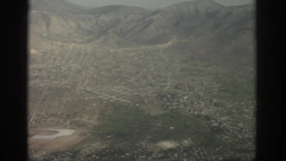 1963 Zurich aerial frame showing neighborhoods, green patches, hills, and a pale airfield area through soft 8mm color.