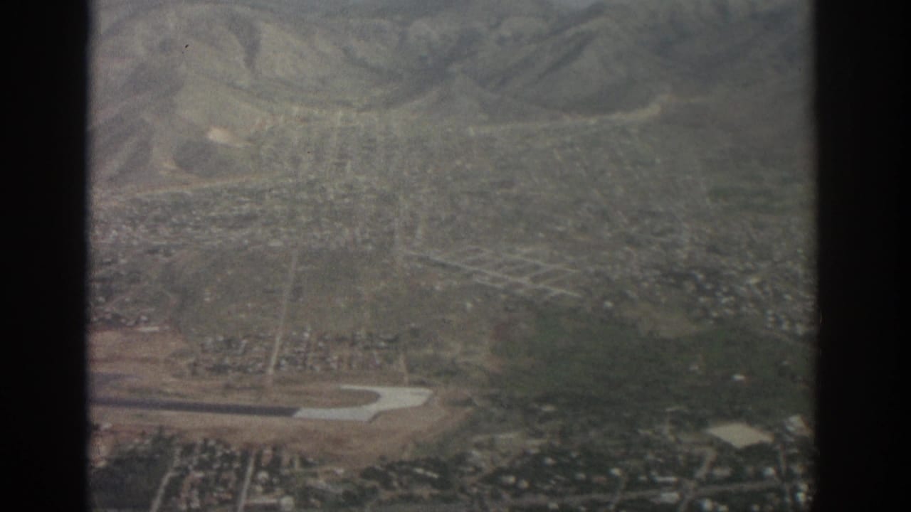 1963 8mm aerial view over Zurich, Switzerland, showing a city grid, runway area, green slopes, and hazy mountains through an aircraft window.
