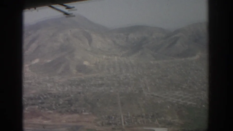1963 8mm aerial footage over Zurich, Switzerland, with a dense city grid below rugged hills and a dark aircraft-window edge at left.
