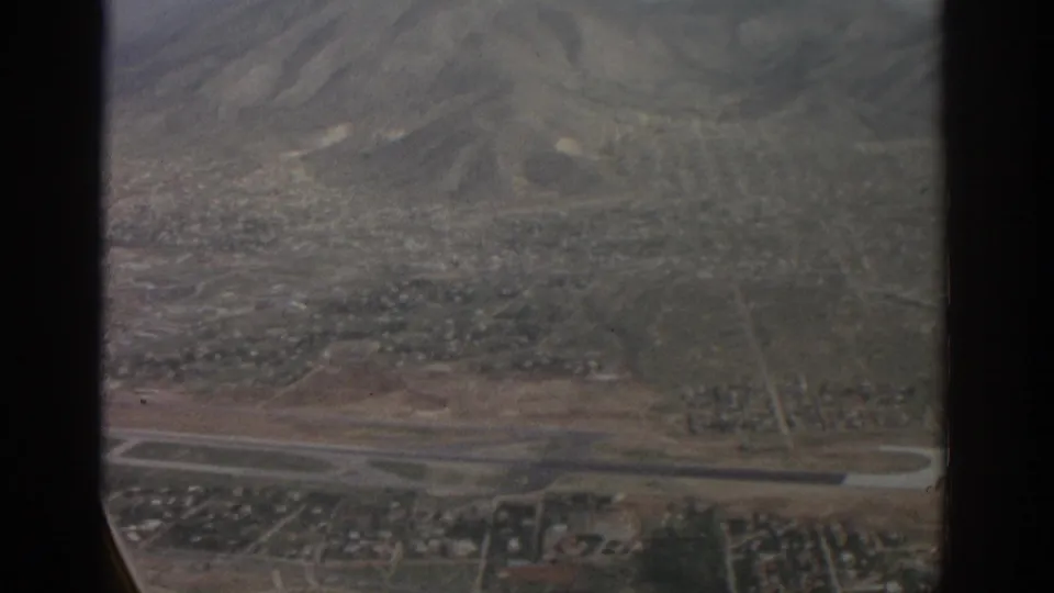 1963 8mm aerial view near Zurich, showing runway infrastructure, neighborhoods, green ground cover, and hazy mountain slopes beyond.