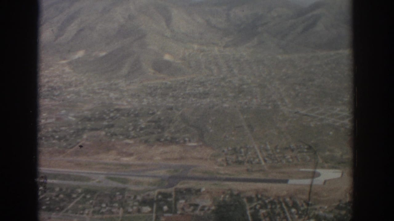 1963 Zurich-area aerial view with low-rise neighborhoods, open fields, a narrow waterway, and mountains in the distance.