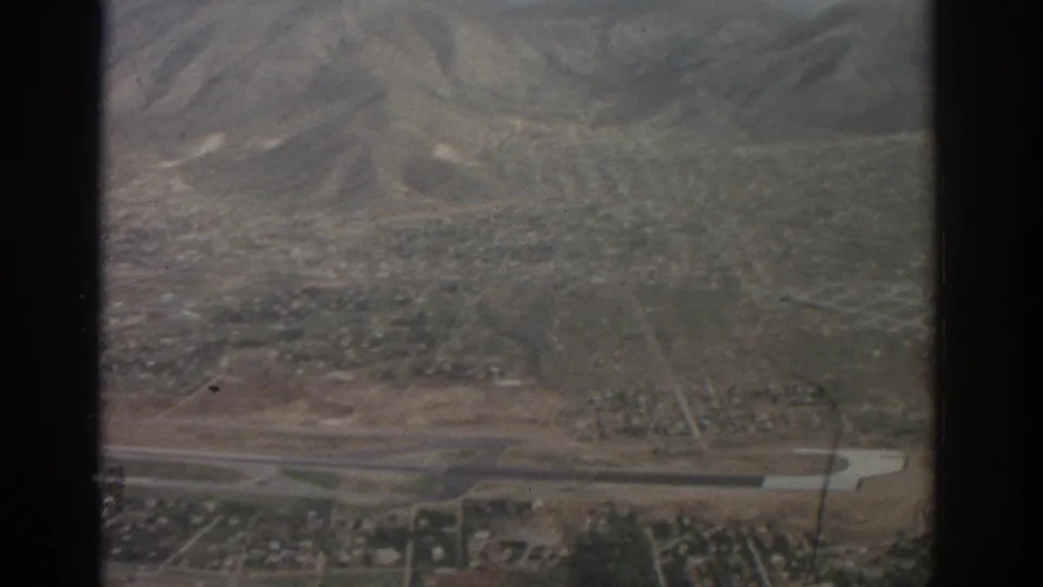 1963 Zurich-area aerial view with low-rise neighborhoods, open fields, a narrow waterway, and mountains in the distance.