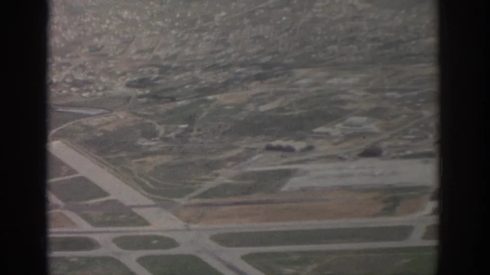 Aerial 8mm view of Zurich-area airport runways, taxiways, open ground, and distant hillside development.