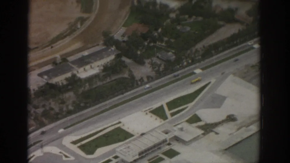 1963 Zurich-area aerial frame showing airport-side roads, buildings, landscaped strips, water, and a small yellow vehicle.