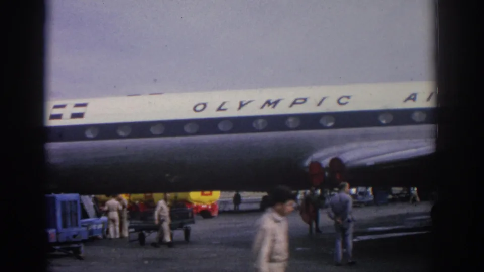 1963 Zurich airport scene with the Olympic Airways fuselage above tarmac vehicles, ground crew and passengers.
