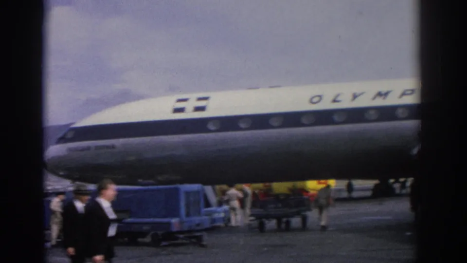 1963 Zurich airport view of the Olympic Airways aircraft nose, blue baggage carts and people crossing the tarmac.