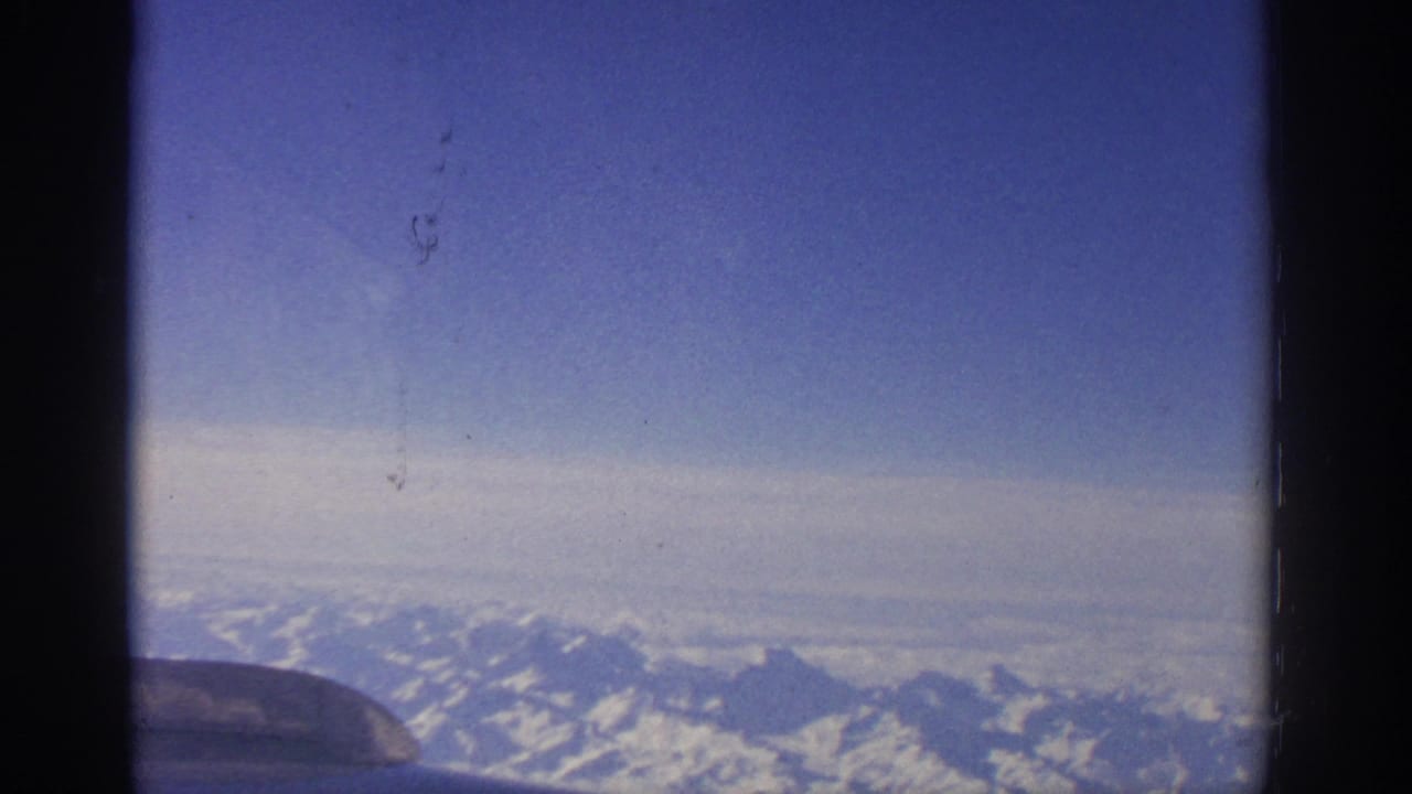 1963 8mm view from an airplane window near Zurich or Switzerland, looking toward snowy alpine ridges beneath a blue sky.