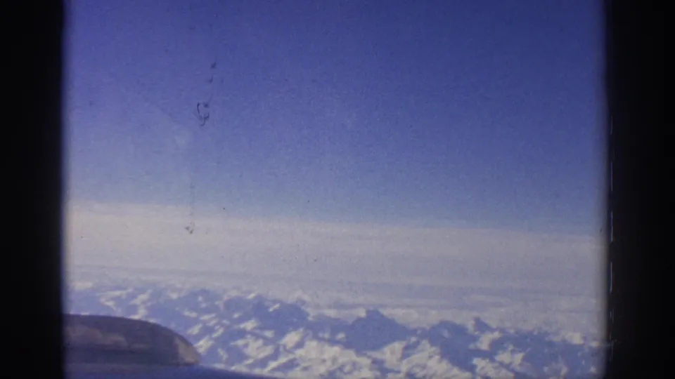 1963 8mm view from an airplane window near Zurich or Switzerland, looking toward snowy alpine ridges beneath a blue sky.