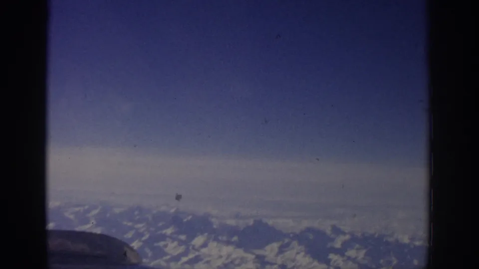 Vintage 8mm airplane-window view from 1963 Switzerland, showing distant snow-covered alpine ridges under blue sky.