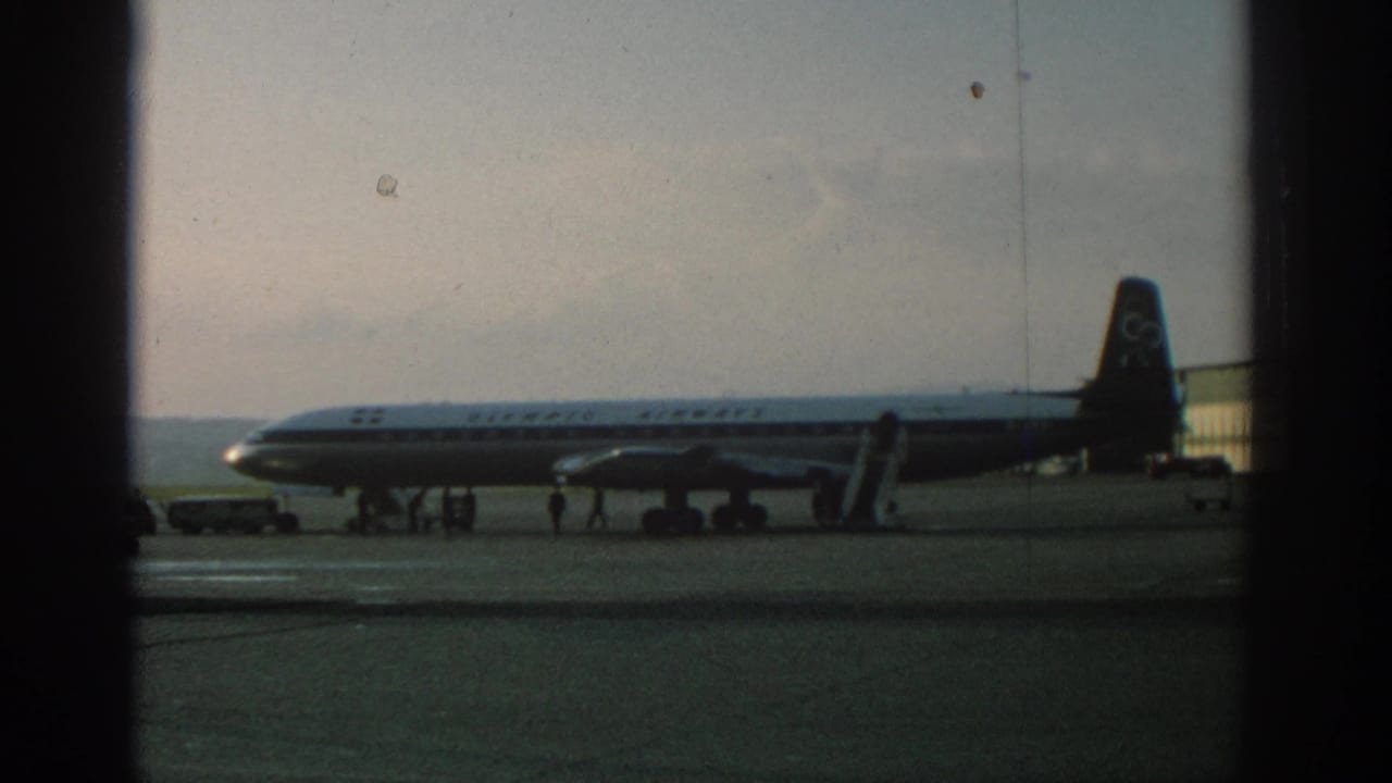 1963 Zurich airport home-movie frame showing an Olympic Airways aircraft parked on the tarmac behind dark frame edges.
