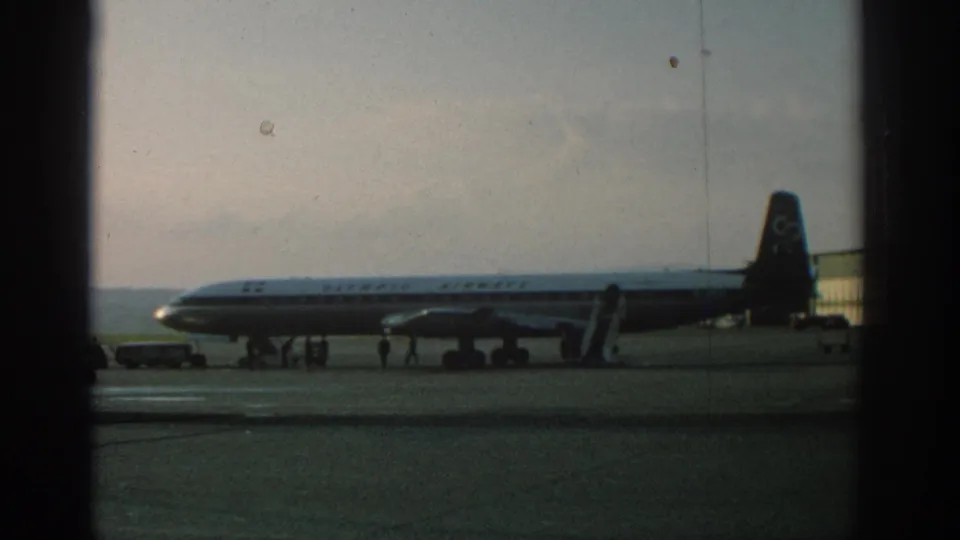 1963 Zurich airport home-movie frame showing an Olympic Airways aircraft parked on the tarmac behind dark frame edges.