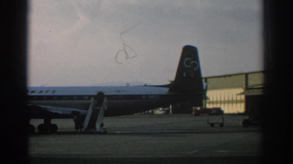 1963 Zurich airport frame showing the rear of an Olympic Airways aircraft beside stairs, terminal glass, and a small service vehicle.