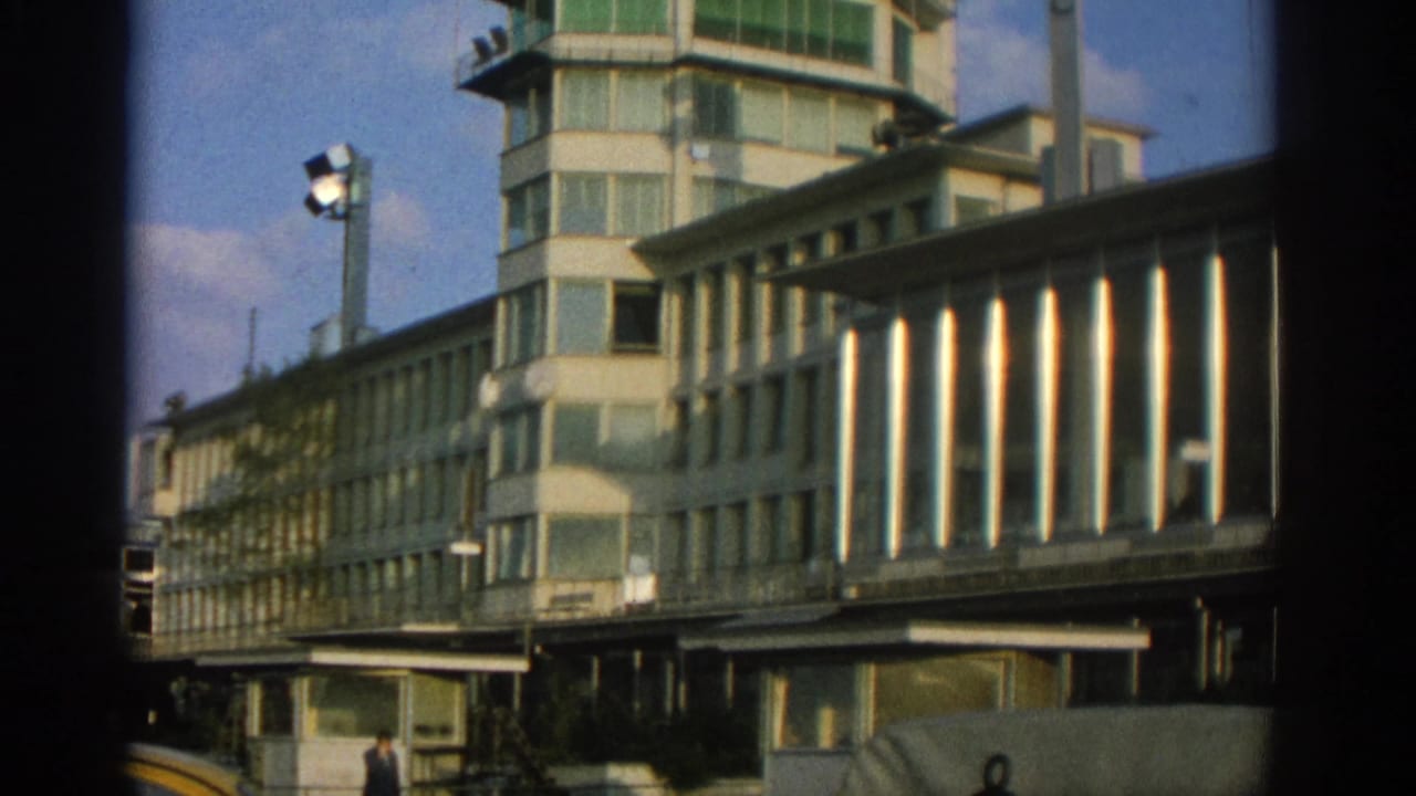 1963 Zurich home-movie frame showing a modernist exterior with green-tinted tower windows, a glazed elevated walkway, and a small pedestrian below.