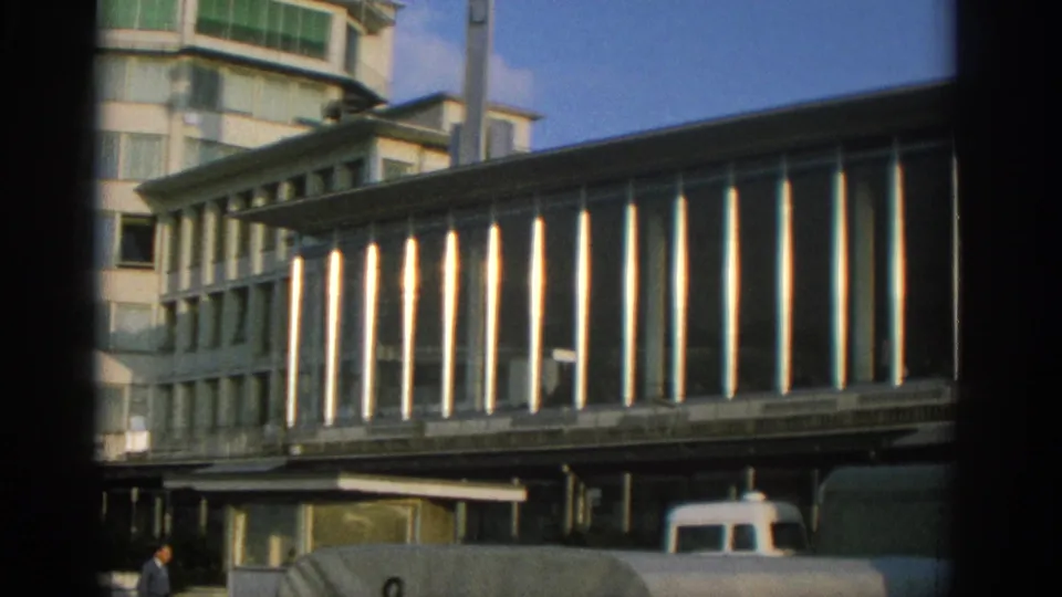1963 Zurich frame showing the elevated glass-panel facade more closely, with vehicles and a small pedestrian near the lower edge.