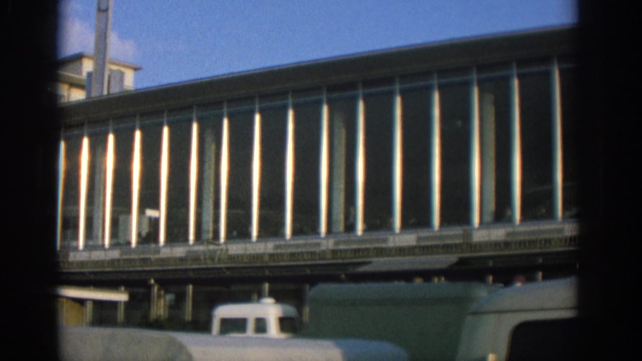 Zurich 1963 street view with a modernist glass facade, bright vertical mullions, and parked vans below.