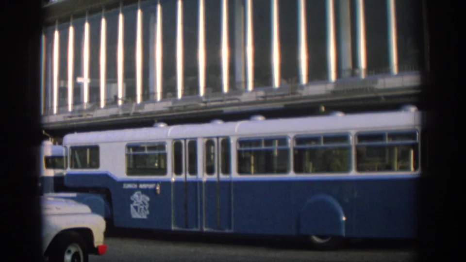 Side view of a blue-and-white Zurich transit vehicle with a pale utility truck in the foreground, filmed on 8mm.