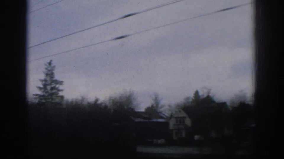 1963 Zurich frame showing small houses partly obscured by trees with overhead wires crossing a cloudy sky.