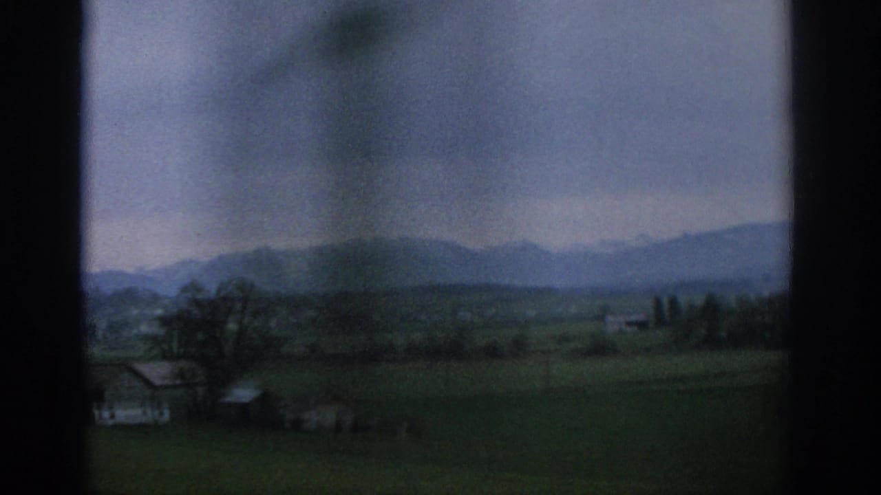 Rural fields and farm buildings seen through dark window framing with mountains in the distance.
